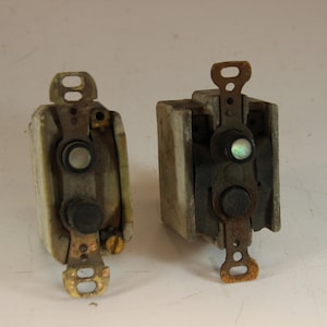 Puede incluir: Dos interruptores eléctricos antiguos. Uno es blanquecino, el otro gris. Ambos tienen componentes metálicos de color latón y botones negros. Los interruptores están montados sobre una superficie blanca.