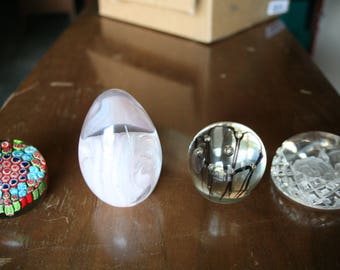 May include: Four decorative glass objects on a brown surface. Includes a colourful mosaic paperweight, an egg-shaped paperweight with white swirls, a clear glass ball with black lines, and a clear, cut-glass paperweight.
