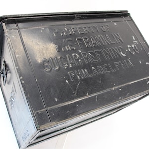 May include: A black metal box with the embossed text "PROPERTY OF THE FRANKLIN SUGAR REFINING CO. PHILADELPHIA." The box has a hinged lid and a latch. The box is slightly angled, showing its rectangular shape.