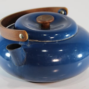 May include: A blue enamel kettle with a wooden handle and lid knob. The kettle has a rounded shape and a spout for pouring. The handle is made of a curved piece of wood attached to the kettle with metal rivets. The lid also has a wooden knob.