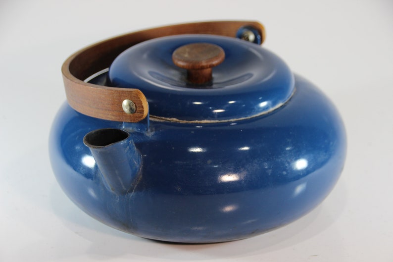 May include: A blue enamel kettle with a wooden handle and lid knob. The kettle has a rounded shape and a spout for pouring. The handle is made of a curved piece of wood attached to the kettle with metal rivets. The lid also has a wooden knob.