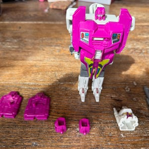 Può includere: Un personaggio d'azione robot rosa, bianco e grigio con più parti staccabili. Il robot ha un grande petto e testa rosa, con gambe grigie e piedi bianchi. Gli accessori includono una pistola grigia e diversi pannelli rosa.