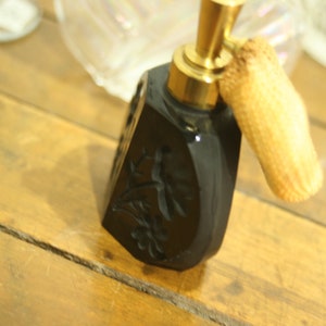 May include: Black perfume bottle with a gold-colored atomizer and a woven puff. The bottle has a floral design carved into the surface. The bottle is on a wooden surface.
