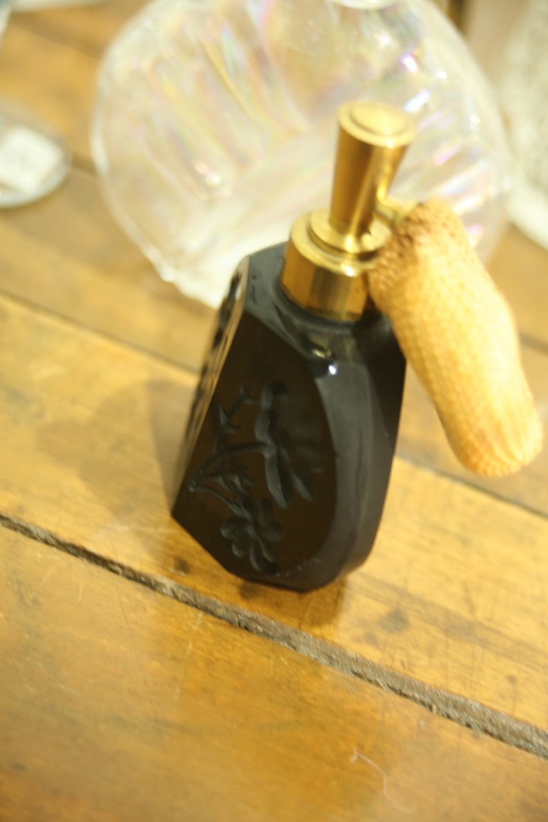 May include: Black perfume bottle with a gold-colored atomizer and a woven puff. The bottle has a floral design carved into the surface. The bottle is on a wooden surface.