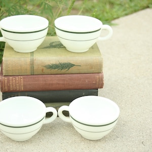 May include: Four white teacups with a dark green stripe detail, arranged on a stack of vintage books. The cups have a classic design with a handle. The books have aged covers with visible titles and textures.
