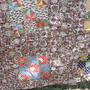 May include: A vintage patchwork quilt with a variety of patterned squares. The quilt features squares with repeating geometric and floral designs in shades of brown, pink, red, and blue. The quilt is hanging outside.
