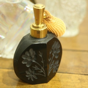 May include: A black perfume bottle with a gold-colored top and a decorative floral design. The bottle has a bulb atomizer with a woven, tan-colored bulb. The bottle is on a wooden surface.