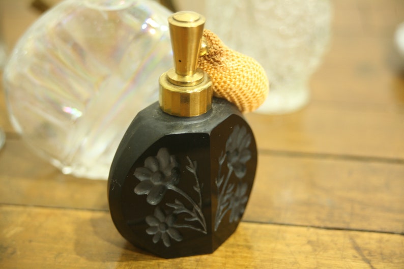 May include: A black perfume bottle with a gold-colored top and a decorative floral design. The bottle has a bulb atomizer with a woven, tan-colored bulb. The bottle is on a wooden surface.