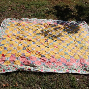 Puede incluir: Un edredón hecho a mano con un diseño de patchwork. El edredón presenta patrones florales amarillos, rosas y azules. El edredón está extendido sobre una superficie cubierta de hierba, mostrando su tamaño y detalles intrincados.