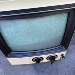Könnte beinhalten: Ein tragbarer Vintage-Fernseher mit cremefarbenem Kunststoffgehäuse und dunkelgrauem Bildschirmrahmen. Die Frontplatte verfügt über zwei große, runde Drehknöpfe und einen kleineren Netzschalter. Der Bildschirm zeigt ein grünes und graues Rauschbild.