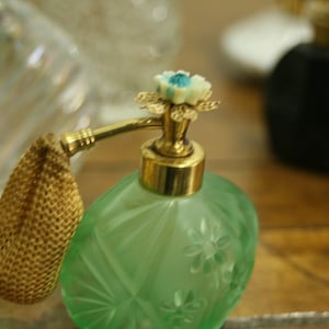 May include: A vintage green perfume bottle with a gold-colored atomizer and floral detailing. The bottle has a textured surface and a decorative flower on top. Other bottles are visible in the background.