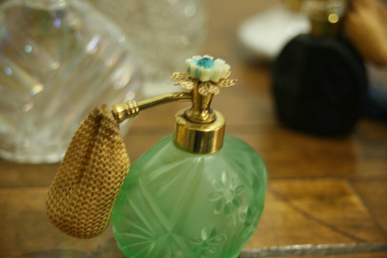 May include: A vintage green perfume bottle with a gold-colored atomizer and floral detailing. The bottle has a textured surface and a decorative flower on top. Other bottles are visible in the background.