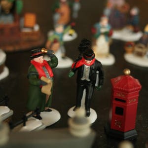 May include: A collection of miniature Christmas village figurines. The figures include a man in a green coat and red scarf, a man in a black coat and top hat, and a red mailbox. The figurines are on a snowy base.