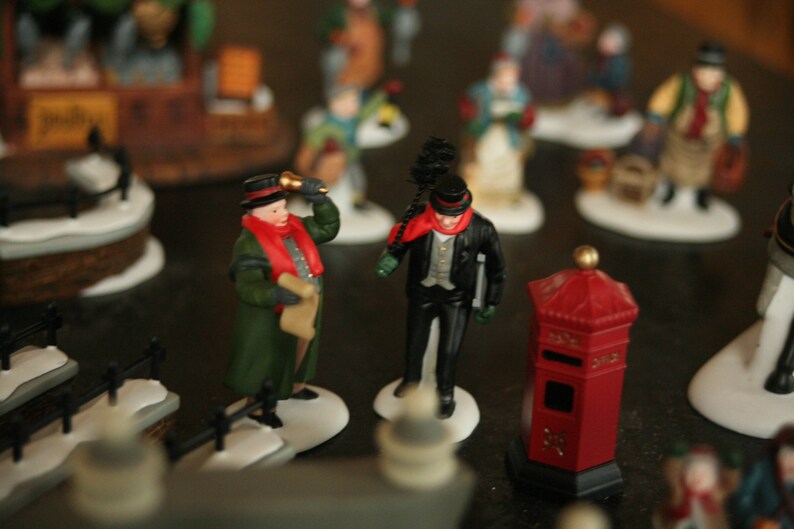 May include: A collection of miniature Christmas village figurines. The figures include a man in a green coat and red scarf, a man in a black coat and top hat, and a red mailbox. The figurines are on a snowy base.