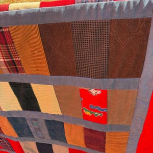 May include: A patchwork quilt with a variety of fabric patterns and textures. The quilt features sections of plaid, corduroy, and patterned fabrics in shades of brown, red, blue, and yellow. The edges are trimmed with a red border.