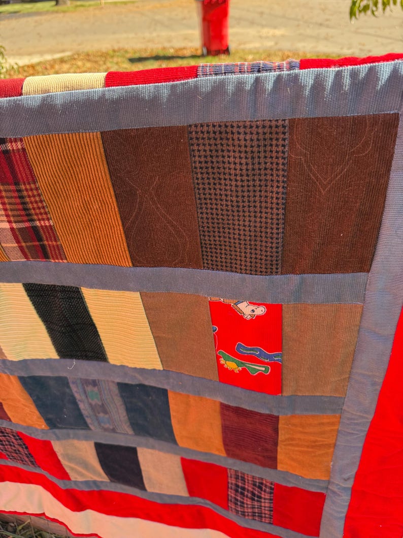 May include: A patchwork quilt with a variety of fabric patterns and textures. The quilt features sections of plaid, corduroy, and patterned fabrics in shades of brown, red, blue, and yellow. The edges are trimmed with a red border.