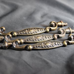 May include: Four antique bronze cabinet pulls. Each pull features a decorative floral design on the center oval and curved ends. The pulls are arranged on a dark gray fabric.