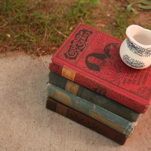 May include: A stack of antique books with red, green, and blue covers, topped with a small white pitcher with a blue floral design. The books are titled "Columbus Series" and "May Bless the Head".