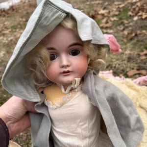 May include: Antique porcelain doll with blonde curly hair, brown eyes, and rosy cheeks. The doll wears a white dress with lace trim and a gray hooded cape. A vintage collectible doll.