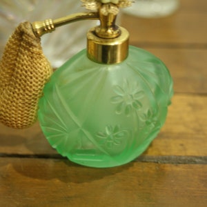May include: A vintage green glass perfume bottle with a gold-colored atomizer and a woven gold-colored bulb. The bottle has a floral design and is sitting on a wooden surface.