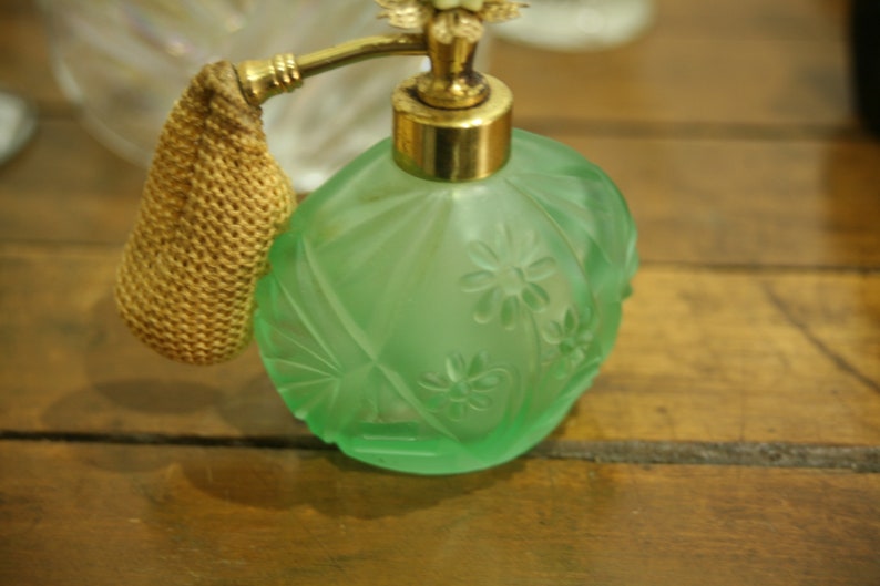 May include: A vintage green glass perfume bottle with a gold-colored atomizer and a woven gold-colored bulb. The bottle has a floral design and is sitting on a wooden surface.