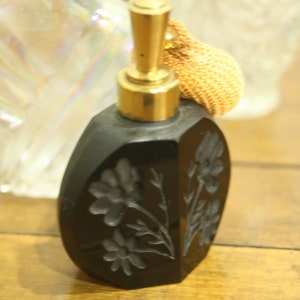 May include: A black perfume bottle with a gold-colored atomizer and a woven bulb. The bottle has a floral design carved into the surface. The bottle is sitting on a wooden surface.