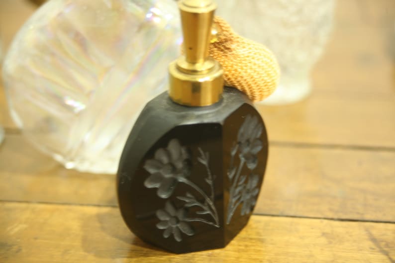 May include: A black perfume bottle with a gold-colored atomizer and a woven bulb. The bottle has a floral design carved into the surface. The bottle is sitting on a wooden surface.