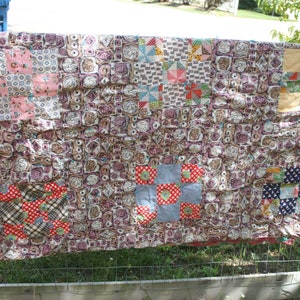 May include: A handmade quilt with a patchwork design, featuring squares of various patterns and colors. The quilt is draped over a wooden fence, showcasing its intricate details and craftsmanship. The color palette includes pink, red, blue, and brown.