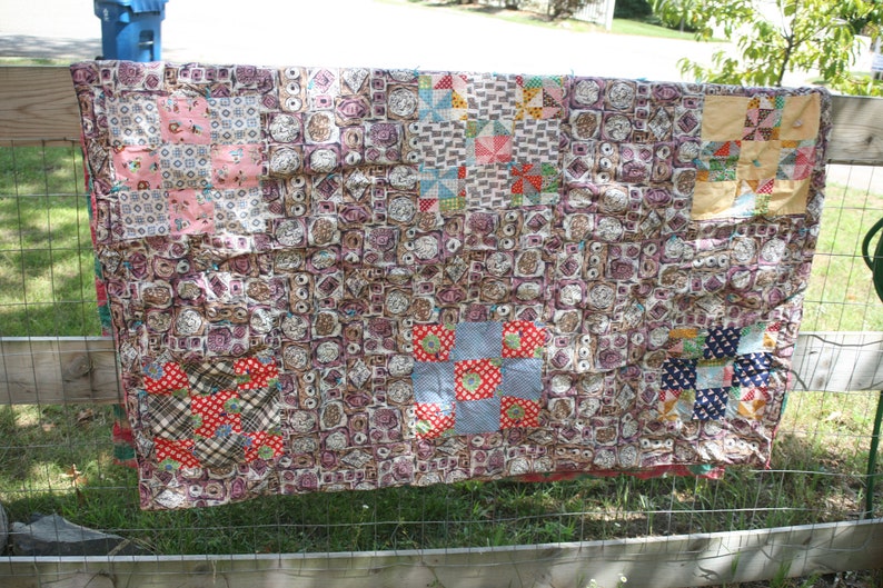 May include: A handmade quilt with a patchwork design, featuring squares of various patterns and colors. The quilt is draped over a wooden fence, showcasing its intricate details and craftsmanship. The color palette includes pink, red, blue, and brown.