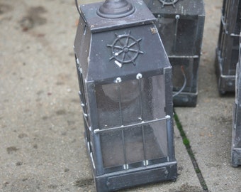May include: Several dark metal lanterns with glass panels and a ship's wheel design on top. The lanterns are rectangular and appear to be vintage or antique. The lanterns are sitting on a concrete surface.