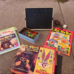 Może przedstawiać: Zestaw zabawek Lite-Brite w stylu vintage z czarnym ekranem, puszką kolorowych kołków i kilkoma pudełkami z wkładami do zdjęć. Pudełka przedstawiają Myszkę Miki Disneya, Królika Bugsa i Pana Główkę. Zestaw znajduje się na betonowej powierzchni.