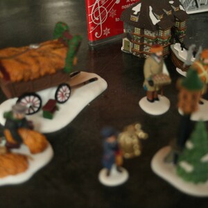 May include: A collection of miniature Christmas village figurines. The scene includes figures of people, a hay wagon, buildings, and trees, all set on snowy bases. The figurines are painted with various colors and details.