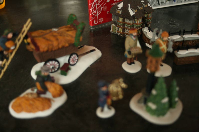 May include: A collection of miniature Christmas village figurines. The scene includes figures of people, a hay wagon, buildings, and trees, all set on snowy bases. The figurines are painted with various colors and details.