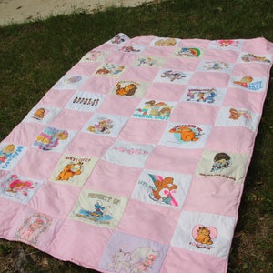 May include: A pink patchwork quilt with cartoon character squares, laid on grass. The quilt features various cartoon characters and phrases like "WORK OUT" and "PROPERTY OF". The quilt is a mix of white and pink squares.