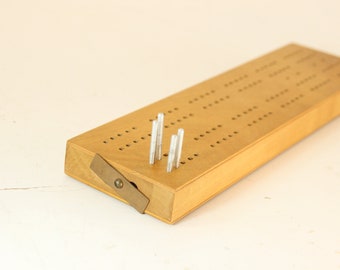 May include: A wooden cribbage board with metal pegs. The board is rectangular with rows of holes for scoring. A metal piece is attached to the side. The board is made of light-coloured wood.