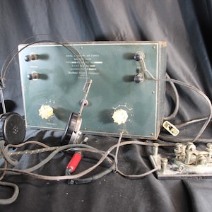 May include: Vintage Royal Canadian Air Force radio equipment. The set includes a green metal box with dials and text, headphones, and a telegraph key. The equipment is connected by wires and cords.