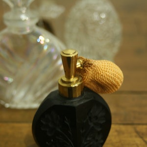 May include: A black perfume bottle with a gold-colored top and a woven atomizer. The bottle has a floral design and is set against a blurred background of other perfume bottles.