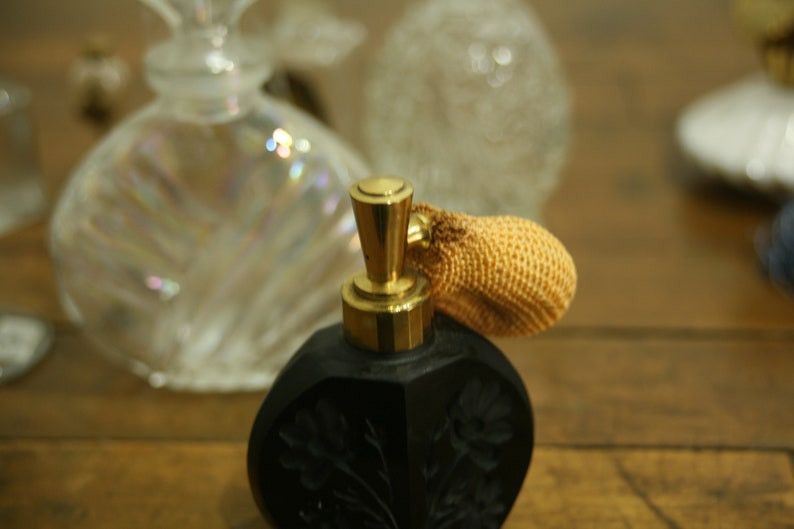 May include: A black perfume bottle with a gold-colored top and a woven atomizer. The bottle has a floral design and is set against a blurred background of other perfume bottles.