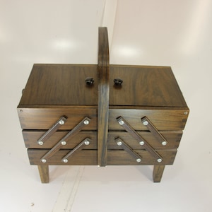 Puede incluir: Una caja de costura de madera marr&oacute;n oscuro con asa y dos peque&ntilde;os pomos. Cuenta con dos cajones extra&iacute;bles con detalles plateados decorativos. La caja se apoya sobre cuatro patas peque&ntilde;as. Aproximadamente 30 cm de altura.