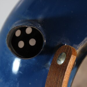May include: Close-up of a blue object with a circular opening featuring four white dots. A wooden handle is attached with a metal fastener. The object has a glossy finish and a curved shape.