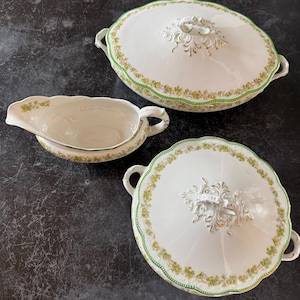 以下が含まれることがあります： 白磁の食器セット。花柄と金色のアクセントが特徴です。蓋付きのサービングディッシュ2つとグレイビーボートが含まれています。縁と端は緑の花柄で装飾されています。蓋には装飾的な白いハンドルが付いています。