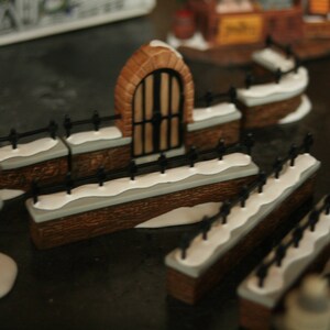 May include: A miniature Christmas village scene featuring a brick archway with a black gate, flanked by snow-covered walls and black fences. The scene includes figurines of people and buildings, creating a festive holiday display.