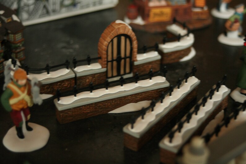 May include: A miniature Christmas village scene featuring a brick archway with a black gate, flanked by snow-covered walls and black fences. The scene includes figurines of people and buildings, creating a festive holiday display.
