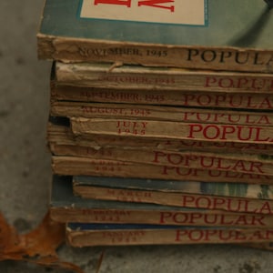 May include: A stack of vintage "Popular Mechanics" magazines from 1945. The magazines have a worn, aged appearance with visible titles and dates, including "November 1945" and "Popular Mechanics" in red text. The covers are a mix of blue and tan.