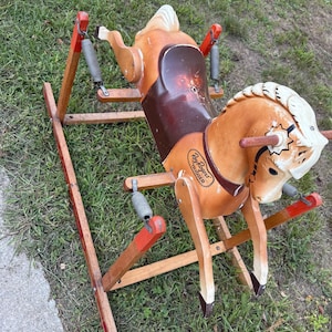 May include: A vintage spring-mounted rocking horse with a tan body, white mane, and a dark brown saddle. The side of the horse features the text Roy Rogers Trigger. It is supported by a wooden frame with red accents and metal springs.