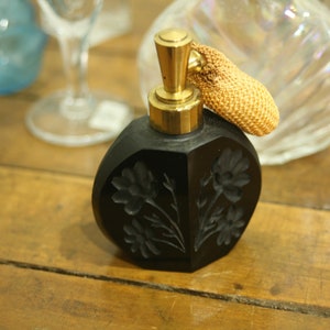 May include: Black perfume bottle with a gold-colored top and a woven atomizer. The bottle has a faceted design with etched floral details. The background includes other glass items on a wooden surface.