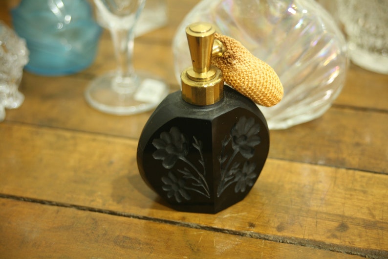May include: Black perfume bottle with a gold-colored top and a woven atomizer. The bottle has a faceted design with etched floral details. The background includes other glass items on a wooden surface.