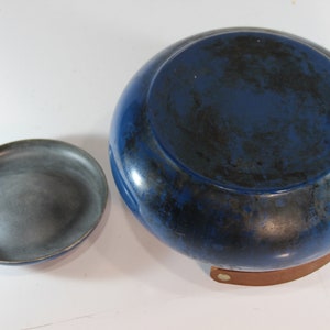 May include: A blue and black ceramic bowl with a matching small dish. The bowl has a rounded shape and a dark blue glaze with black accents. The small dish is gray. The bowl sits on a brown leather base.