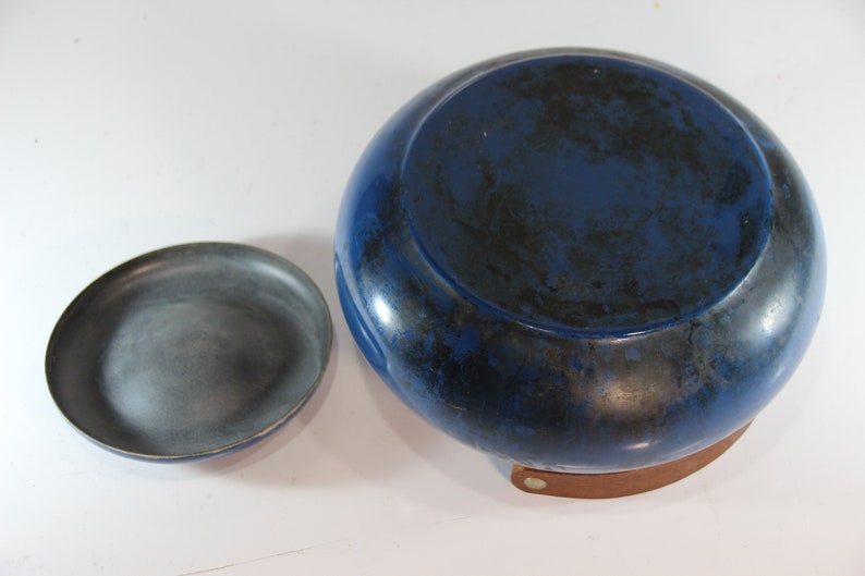 May include: A blue and black ceramic bowl with a matching small dish. The bowl has a rounded shape and a dark blue glaze with black accents. The small dish is gray. The bowl sits on a brown leather base.