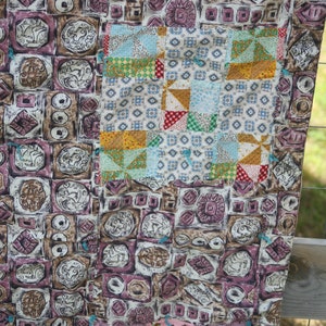 May include: A patchwork quilt with a mix of patterned fabrics. The main section features repeating square designs in brown, purple, and cream. A smaller section has colorful squares and triangles. The quilt is a mix of vintage and modern styles.
